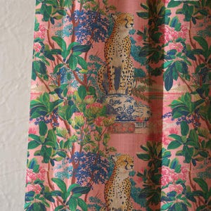 May include: Pink fabric with a repeating pattern of cheetahs, foliage, and floral elements. The cheetahs are depicted with their distinctive spots, and the design includes green leaves, blue accents, and pink flowers. The fabric has a textured appearance.