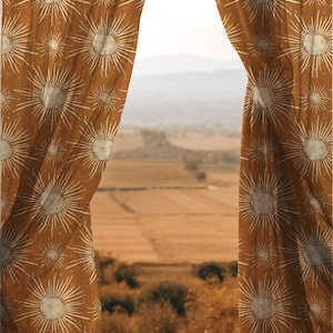 May include: Brown curtains, patterned with white sunbursts, are open to reveal a rural vista. The curtains' design contrasts with the fields, trees, and distant hills. The colour scheme is warm, featuring earthy tones.