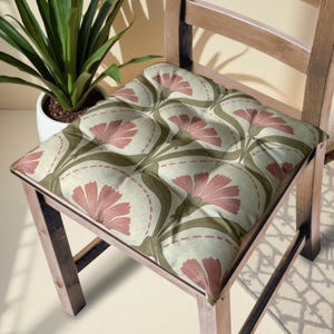 May include: A wooden chair with a floral cushion. The cushion features a repeating pattern of pink flowers and green stems on a cream background. The chair is made of light-colored wood and has a simple design. A potted plant is visible in the background.