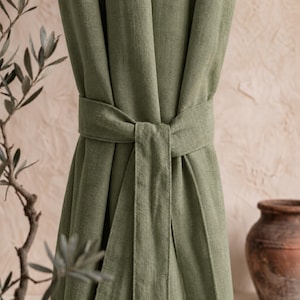 Peut inclure: Rideau en lin vert olive noué avec une ceinture en tissu assortie. Le rideau est drapé et froncé, avec une texture visible. Une branche d'olivier et un vase en terre cuite sont en arrière-plan.