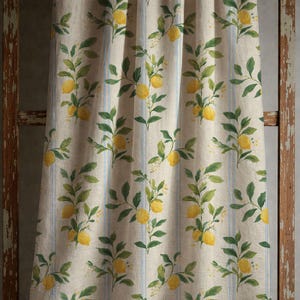 May include: A beige fabric curtain featuring a repeating pattern of yellow lemons and green leaves on branches, accented by vertical light blue stripes. The textile has a visible woven texture and hangs in soft, vertical folds across the surface.