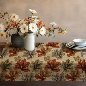 May include: A beige table runner featuring a repeating pattern of autumn leaves in brown, orange, and grey. The runner is on a wooden table, with a vase of flowers and a set of plates and bowls.