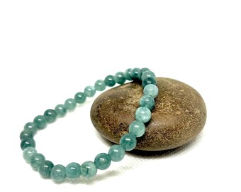 May include: A green gemstone bracelet with round beads, resting on a brown rock.