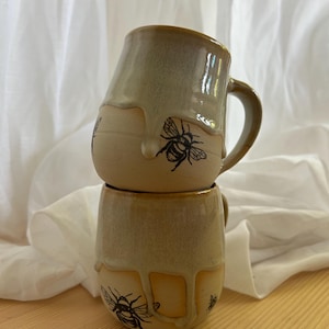 Może przedstawiać: Dwa ceramiczne kubki ułożone w stos z beżową glazurą i brązową obwódką. Każdy kubek ma czarną ilustrację pszczoły i wzór z kroplami glazury. Kubki są ułożone na drewnianej powierzchni.