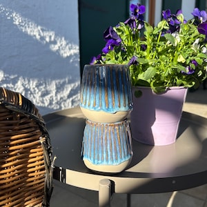 Może przedstawiać: Dwa ułożone na sobie niebieskie i brązowe kubki ceramiczne z fakturowaną glazurą. Kubki stoją na szarym stole z wiklinowym krzesłem w tle.