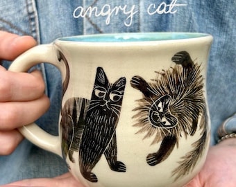 May include: A cream-colored ceramic mug with a light blue interior, featuring black ink illustrations of two cats. One cat stands calmly while the other appears agitated with spiky fur. The text "angry cat" is written in white script above the cats.