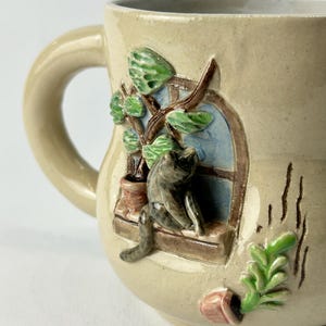 Op de afbeelding: Beige keramische mok met een gebeeldhouwde kat in een raam met een plant. De mok heeft een gebogen handvat. Het raam heeft een blauwe achtergrond.