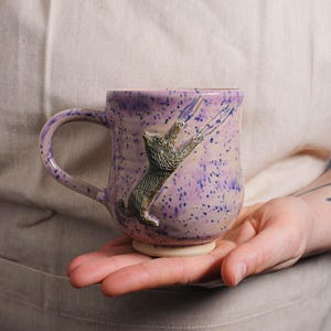 May include: A purple ceramic mug with a 3D cat design. The cat is climbing up the side of the mug and has a speckled purple glaze.