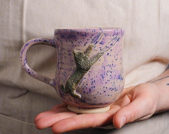 May include: A purple ceramic mug with a 3D cat design. The cat is climbing up the side of the mug and has a speckled purple glaze.