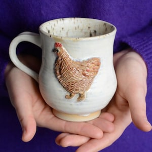 Può includere: Una tazza in ceramica bianca con un design di pollo marrone e rosso. La tazza ha una glassa macchiata e un manico.