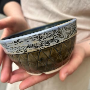 Pode incluir: Uma tigela de cerâmica artesanal com um desenho de dragão. A tigela apresenta um padrão de diamante na metade inferior e uma borda azul clara com uma ilustração de dragão preto. A tigela é segurada nas mãos de uma pessoa.