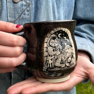 Może przedstawiać: Ciemnobrązowy ceramiczny kubek z kremową podstawą. Kubek posiada szczegółową kremową ilustrację stworzenia podobnego do kota pod półksiężycem. Kubek ma zakrzywiony uchwyt i jest trzymany przez osobę.