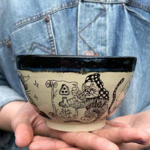Może przedstawiać: Ceramiczna miska z kremową podstawą i ciemnoniebieskim brzegiem. Zewnętrzna strona ozdobiona jest czarnymi rysunkami liniowymi przedstawiającymi kota w czapce w szachownicę, trzymającego zwój z trójkątnym symbolem, w otoczeniu gwiazdek i roślin.