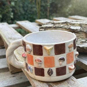 Op de afbeelding: Een keramische mok met een crèmekleurige basis en handvat. De mok heeft een dambordpatroon van bruine en oranje vierkanten, versierd met 3D-paddenstoelontwerpen. De handgreep heeft een getextureerde, organische vorm.
