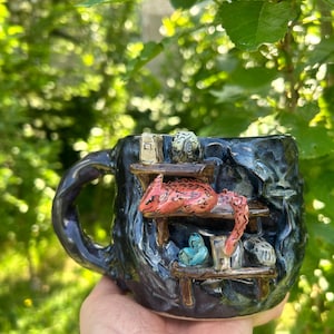 Könnte beinhalten: Ein dunkelblauer, texturierter Keramikbecher mit einer skulptierten Innenszene und zwei holzartigen Regalen. Eine rote, gefleckte Kreatur ruht auf dem oberen Regal, während kleine blaue und weiße Objekte auf dem unteren Regal vor einer dunklen Wand stehen.