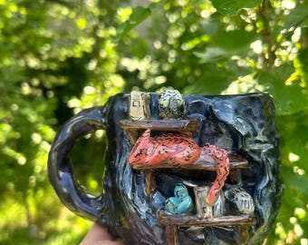 Può includere: Una tazza in ceramica blu scuro testurizzata con una scena interna scolpita e due ripiani in legno. Una creatura rossa maculata riposa sul ripiano superiore, mentre piccoli oggetti blu e bianchi si trovano su quello inferiore contro una parete scura.