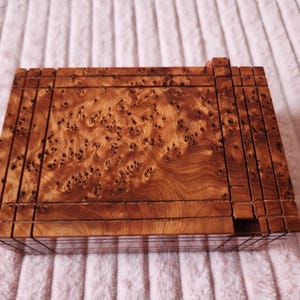Könnte beinhalten: Eine rechteckige Holzkiste mit einem natürlichen Maserungsmuster. Die Kiste ist aus einer Reihe vertikaler und horizontaler Holzleisten gefertigt, wodurch eine strukturierte Oberfläche entsteht. Das Holz hat einen warmen, rötlich-braunen Farbton.