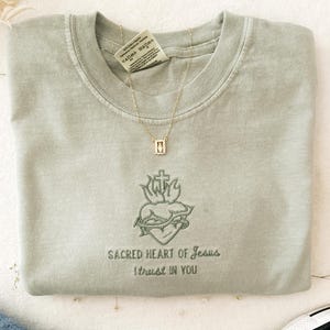以下が含まれることがあります： セージグリーンのTシャツに、棘と炎がついた心臓の刺繍が施されています。心臓の下には「Sacred Heart of Jesus I trust in you」という刺繍が施されています。