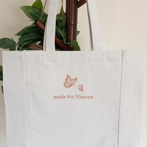 May include: Off-white canvas tote bag with two butterflies and the text "made for Heaven" in brown. The bag has long handles and is hanging on a wooden rack. The background includes green leaves.