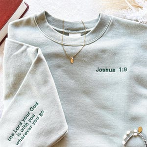 Pode incluir: Camisola verde-sálvia com o texto "the Lord your God is with you wherever you go" na manga esquerda e "Joshua 1:9" no lado direito do peito.