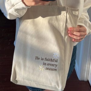 May include: A cream-coloured tote bag featuring embroidered text and floral details. The text says "He is faithful in every season." The bag is held by someone wearing a white sweatshirt.