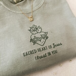 以下が含まれることがあります： 薄い緑色のTシャツに、十字架と棘のある心臓の刺繍が施されています。心臓の下には「SACRED HEART OF JESUS I TRUST IN YOU」という刺繍が施されています。