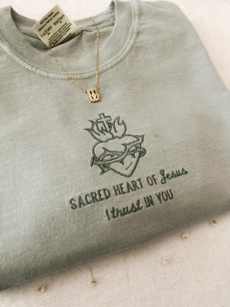 以下が含まれることがあります： 薄い緑色のTシャツに、十字架と棘のある心臓の刺繍が施されています。心臓の下には「SACRED HEART OF JESUS I TRUST IN YOU」という刺繍が施されています。