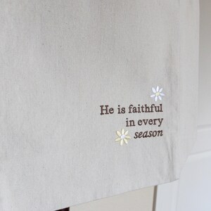 May include: Beige tote bag with embroidered text and floral details. The text reads "He is faithful in every season." Features two small daisy designs, one white and one yellow.