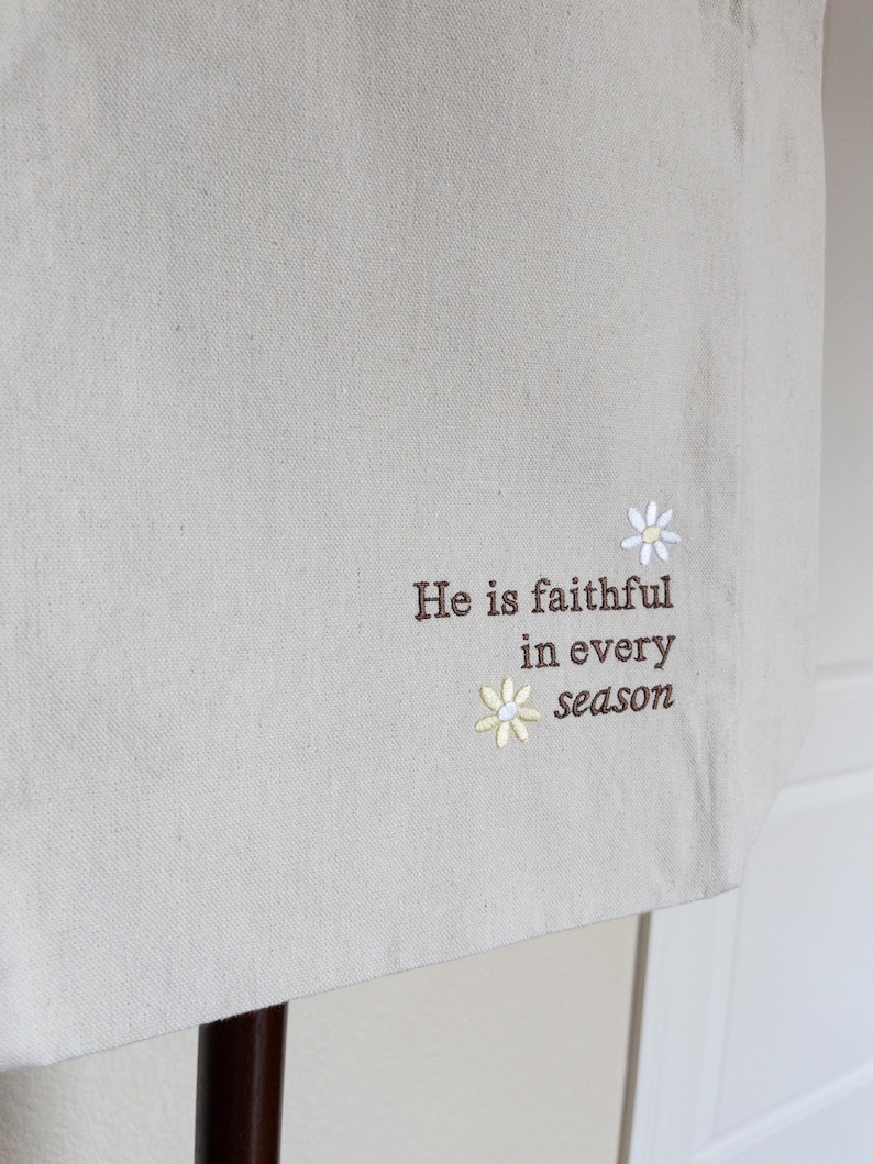 May include: Beige tote bag with embroidered text and floral details. The text reads "He is faithful in every season." Features two small daisy designs, one white and one yellow.