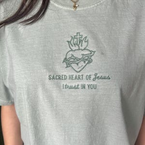 以下が含まれることがあります： 薄い緑色のTシャツに、十字架、棘、炎がついた心臓の刺繍が施されています。 「Sacred Heart of Jesus I trust in you」という文字が、画像の下に刺繍されています。