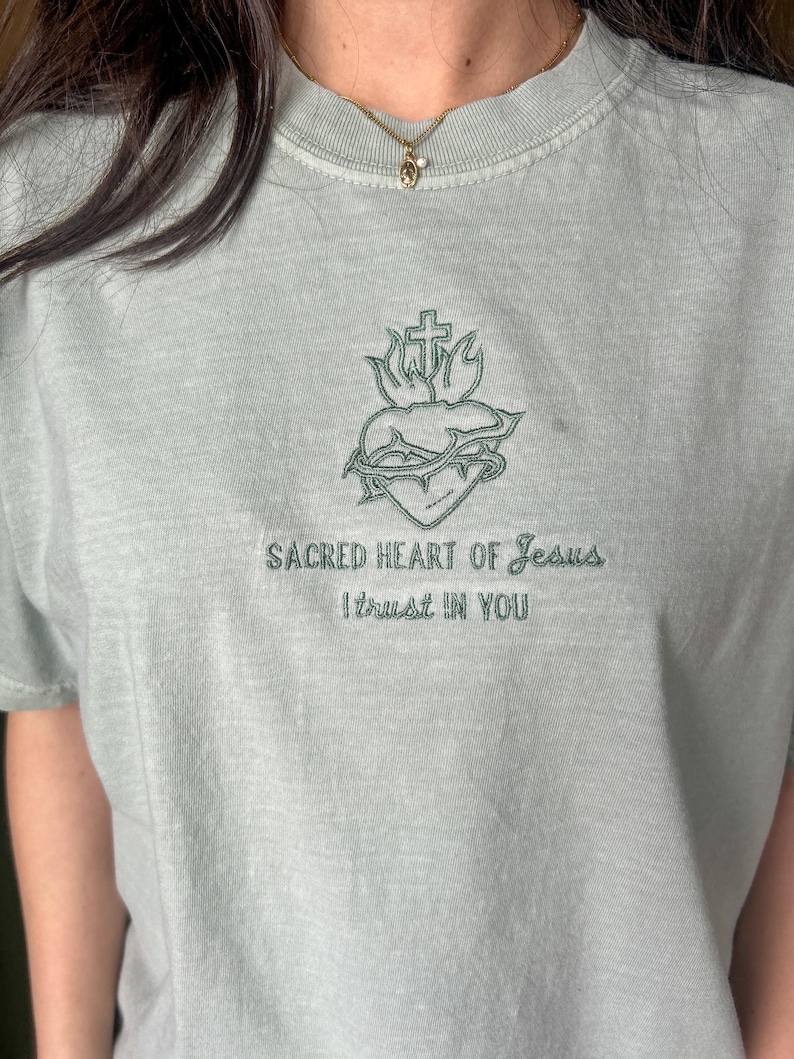 以下が含まれることがあります： 薄い緑色のTシャツに、十字架、棘、炎がついた心臓の刺繍が施されています。 「Sacred Heart of Jesus I trust in you」という文字が、画像の下に刺繍されています。