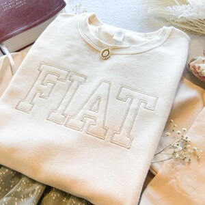 Pu&ograve; includere: Una felpa color crema con la parola "FIAT" ricamata in un grande e audace font. La felpa &egrave; piegata e mostrata su una superficie bianca con un libro, fiori e gioielli.