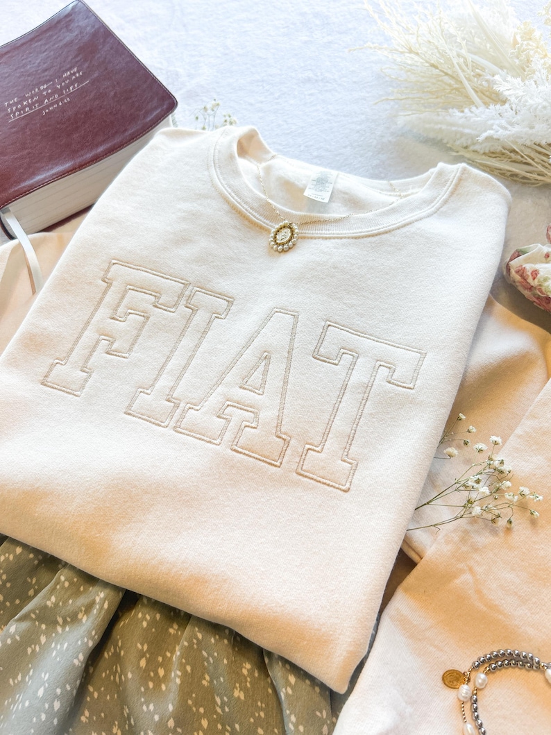 Pu&ograve; includere: Una felpa color crema con la parola "FIAT" ricamata in un grande e audace font. La felpa &egrave; piegata e mostrata su una superficie bianca con un libro, fiori e gioielli.