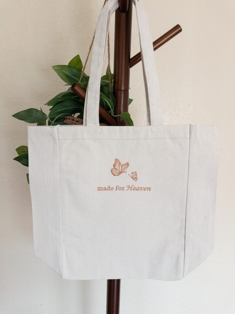 May include: Off-white canvas tote bag with two butterflies and the text "made for Heaven" in brown. The bag has long handles and is hanging on a wooden rack. The background includes green leaves.