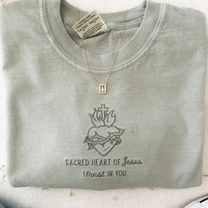 以下が含まれることがあります： セージグリーンのTシャツに、十字架、棘、炎がついた心臓の刺繍が施されています。 「SACRED HEART OF JESUS I TRUST IN YOU」という文字が、画像の下に刺繍されています。