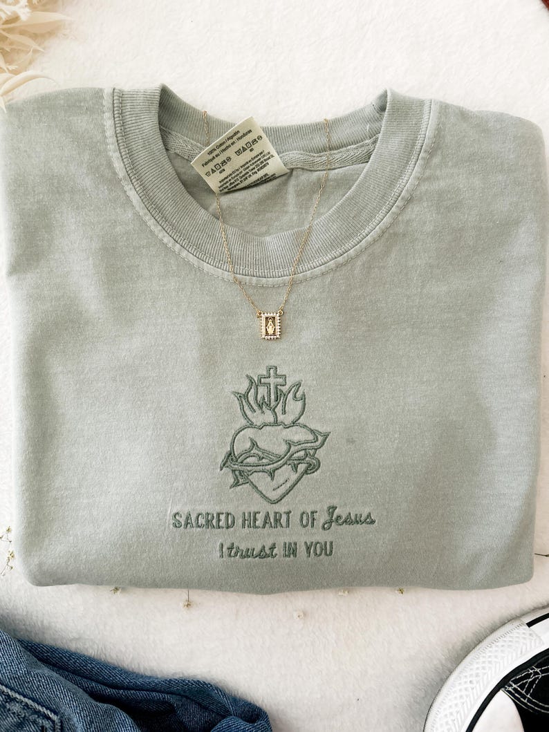 以下が含まれることがあります： セージグリーンのTシャツに、十字架、棘、炎がついた心臓の刺繍が施されています。 「SACRED HEART OF JESUS I TRUST IN YOU」という文字が、画像の下に刺繍されています。