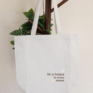 May include: Cream-coloured canvas tote bag with white handles. The bag has the text "He is faithful in every season" in brown script, with a small floral design. The bag is hanging on a wooden coat rack with green leaves.