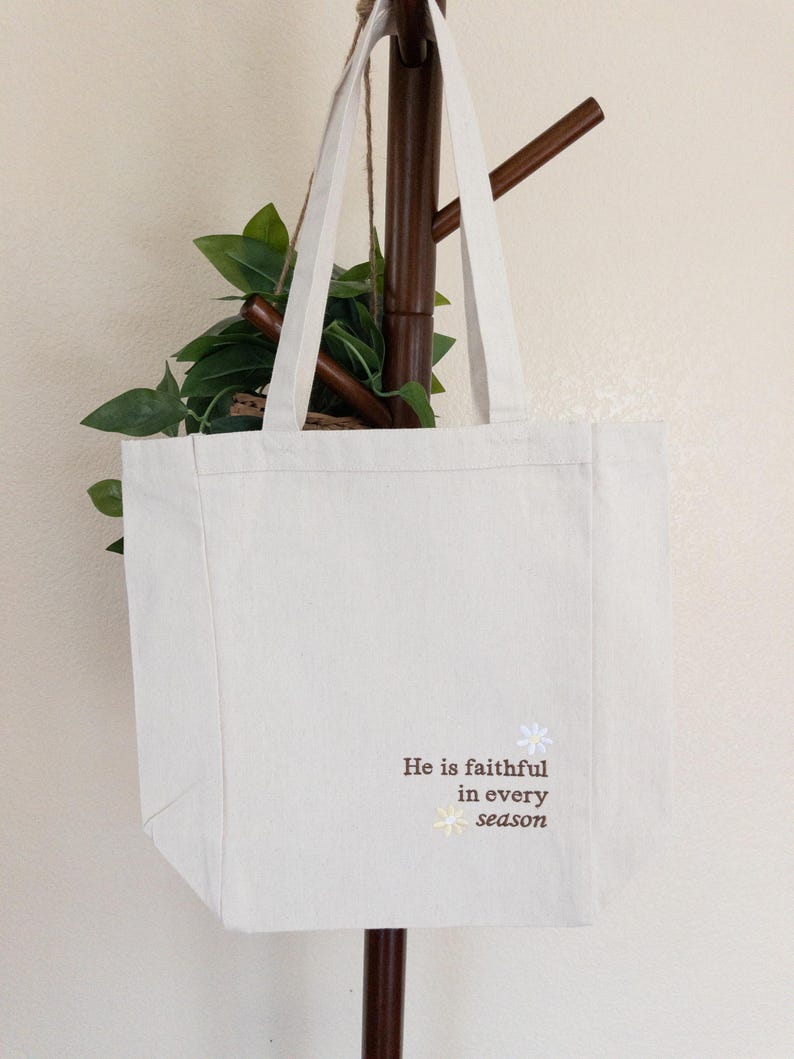 May include: Cream-coloured canvas tote bag with white handles. The bag has the text "He is faithful in every season" in brown script, with a small floral design. The bag is hanging on a wooden coat rack with green leaves.