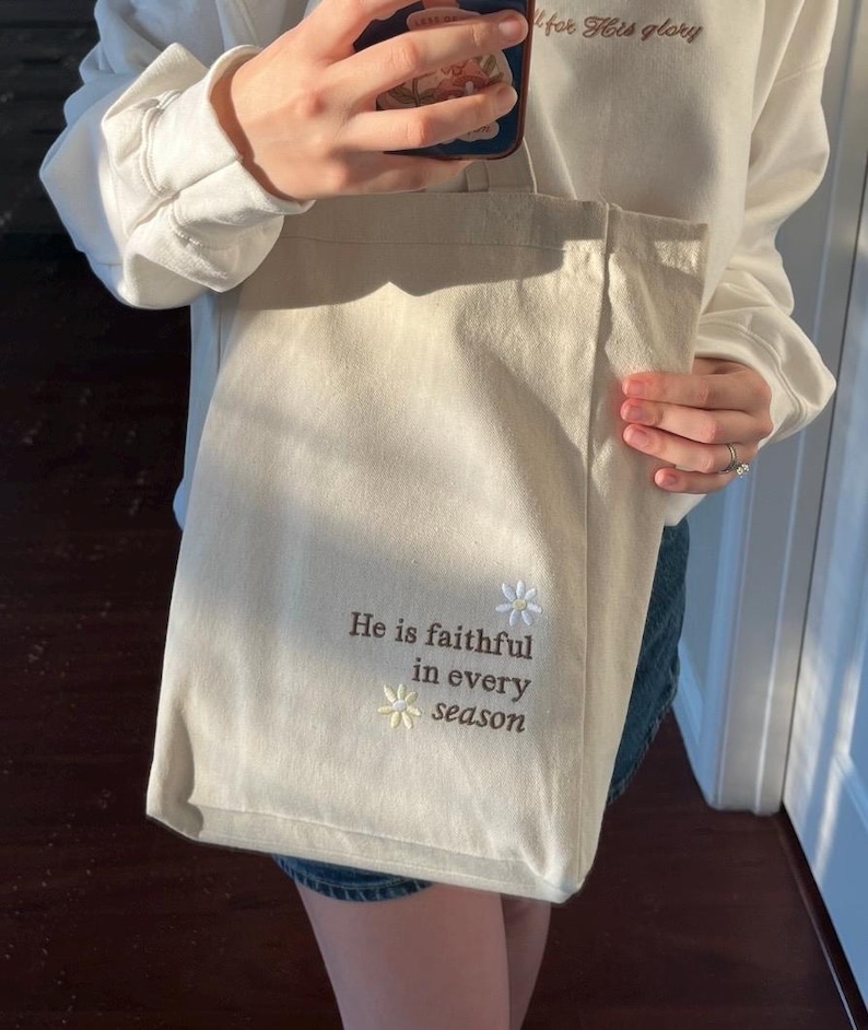May include: A cream-coloured tote bag featuring embroidered text and floral details. The text says "He is faithful in every season." The bag is held by someone wearing a white sweatshirt.