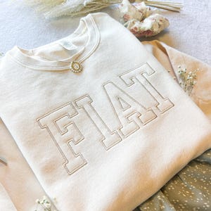 Pu&ograve; includere: Una felpa color crema con la parola "FIAT" ricamata in un grande e audace font. La felpa &egrave; piegata e appoggiata su un tessuto a fantasia.