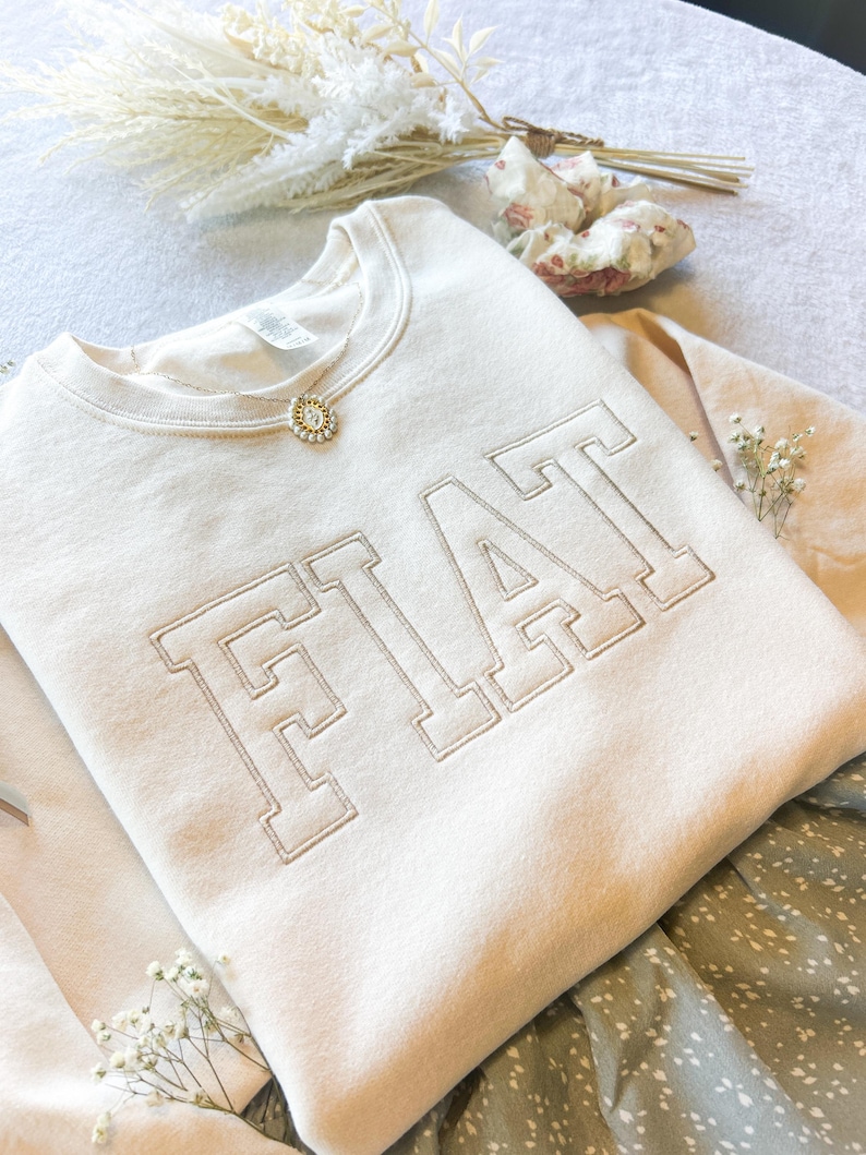 Pu&ograve; includere: Una felpa color crema con la parola "FIAT" ricamata in un grande e audace font. La felpa &egrave; piegata e appoggiata su un tessuto a fantasia.