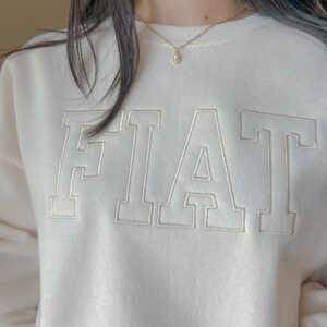 Pu&ograve; includere: Una felpa bianca con la parola "FIAT" ricamata in bianco sul davanti. La felpa ha un collo rotondo e maniche lunghe. La persona che indossa la felpa indossa anche una collana a catena d'oro con un piccolo ciondolo.