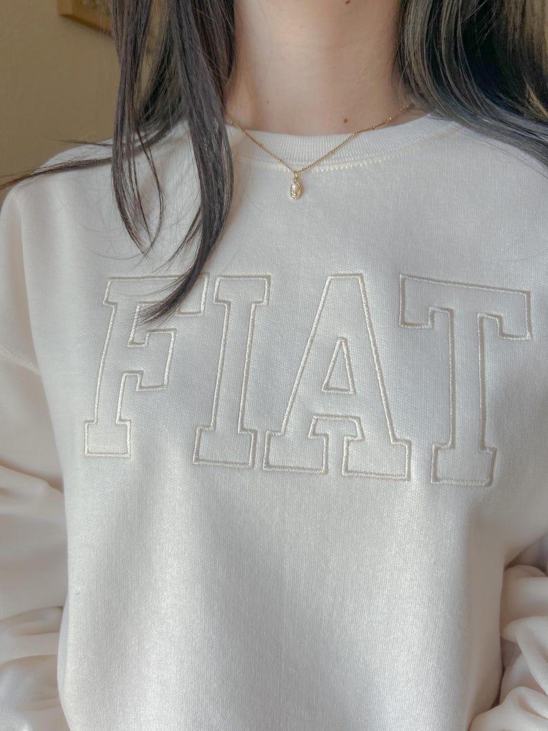 Pu&ograve; includere: Una felpa bianca con la parola "FIAT" ricamata in bianco sul davanti. La felpa ha un collo rotondo e maniche lunghe. La persona che indossa la felpa indossa anche una collana a catena d'oro con un piccolo ciondolo.