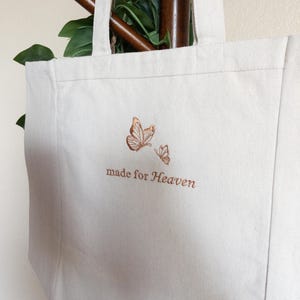 May include: Off-white canvas tote bag with two embroidered butterflies in shades of brown. The words "made for Heaven" are embroidered below the butterflies. The bag has two handles and is hanging on a wooden rod.