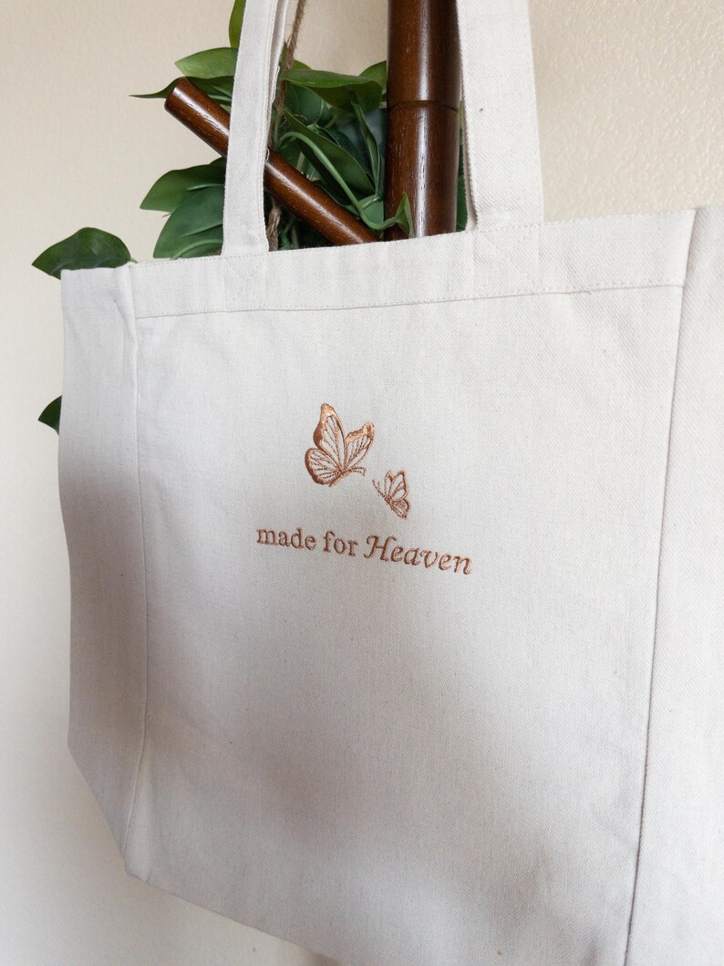 May include: Off-white canvas tote bag with two embroidered butterflies in shades of brown. The words "made for Heaven" are embroidered below the butterflies. The bag has two handles and is hanging on a wooden rod.