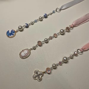 Op de afbeelding: Drie delicate, met kralen bezette lint-bladwijzers met verschillende bedels. De eerste bladwijzer heeft een bedel van blauw-wit porselein, de tweede heeft een cameo-bedel en de derde heeft een witte bloemenbedel.