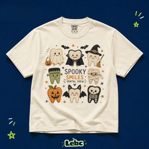 Puede incluir: Una camiseta de manga corta color crema con un gráfico de varios dientes disfrazados para Halloween, incluyendo un fantasma, vampiro, bruja, momia y esqueleto, con el texto "SPOOKY SMILES DENTAL CREW" impreso en el centro.