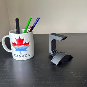 Puede incluir: Un soporte para auriculares de impresión 3D negro con una base curva. El soporte está sobre una superficie de madera oscura. Una taza blanca con un diseño de bandera canadiense y lápices está en el fondo.