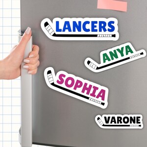 Op de afbeelding: Vier hockey-thema stickers op een koelkast. De stickers tonen namen en het woord "Lancers" in vette, kleurrijke tekst, elk gecombineerd met een hockeystick-afbeelding. De namen zijn Anya, Sophia en Varone.