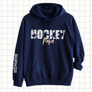 May include: Dark blue hoodie with a front pocket and a drawstring hood. The front displays "HOCKEY Papa" in white and tan, with hockey player silhouettes. The left sleeve has the name "SOPHIA" in white.